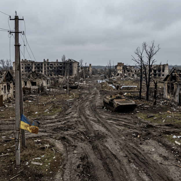Ukrainian flag, abandoned tank, and destroyed buildings along muddy road